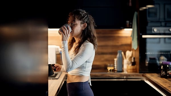 A woman stands in a kitchen, holding a cup of coffee. She enjoys a quiet moment while looking out. The kitchen has wooden cabinets and a