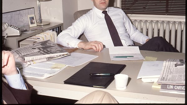Man in tie sits at desk covered with papers/documents, another man sits mostly off camera to left