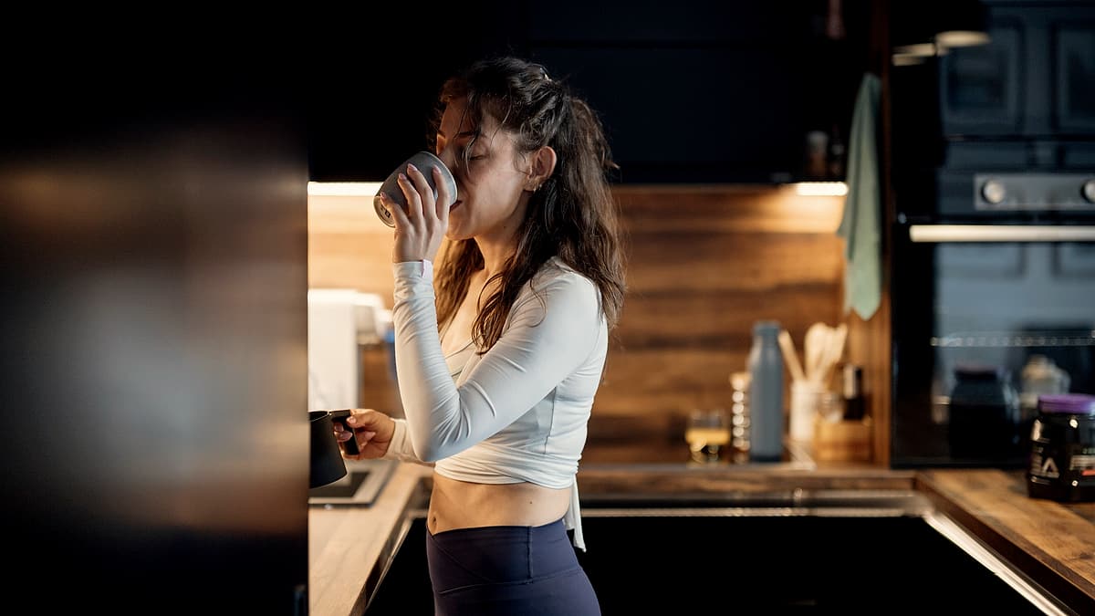 A woman stands in a kitchen, holding a cup of coffee. She enjoys a quiet moment while looking out. The kitchen has wooden cabinets and a
